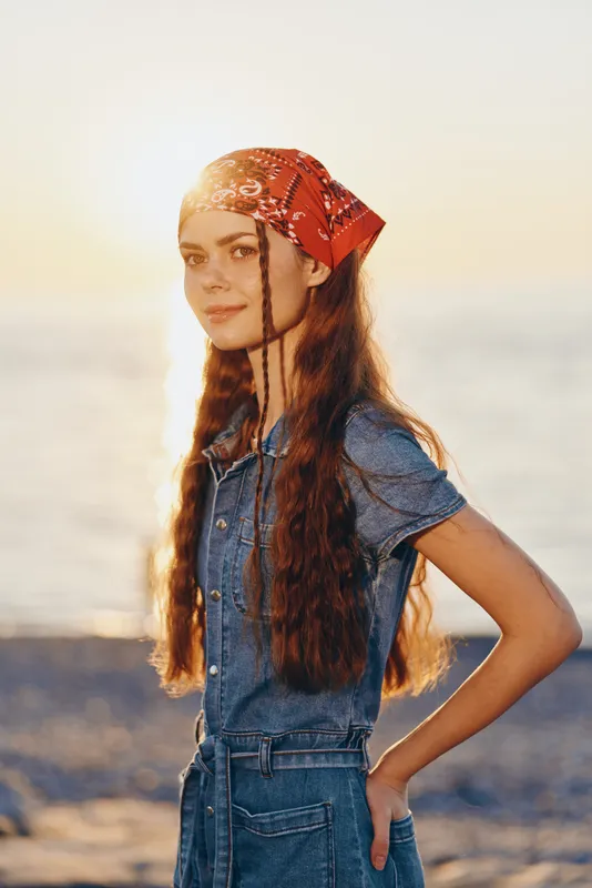 Lifestyle woman in modern boho-western denim street style with long braided hair and red bandana by the beach at sunset, film color warm tones.