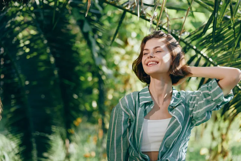 Young woman enjoying sunlight outdoors, smiling with a striped green shirt and white top, surrounded by lush tropical leaves.