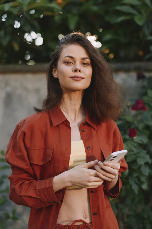 Confident woman outdoors, casual fashion, red shirt, sunlit moment, relaxed pose, gentle breeze, natural colors, lush garden background, soft lighting enhances textures and mood