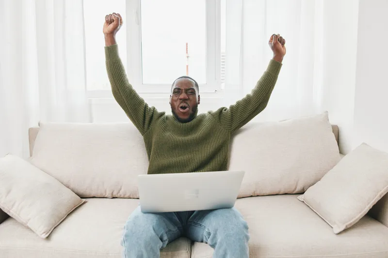 Celebrating success, a Black man in a green sweater joyfully raises his arms on a beige couch with a laptop, showcasing emotions of victory and excitement