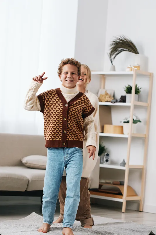 Authentic playful scene shows a child wearing a knitted vest and cozy sweater, ready for school adventures in a bright living space at home.