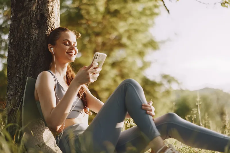 Young woman relaxing outdoors in activewear, listening to music on her phone in nature, enjoying sunshine and a peaceful atmosphere