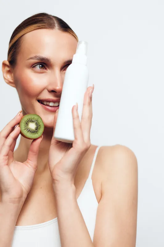 Young woman with clear skin holding a white cosmetic bottle and a sliced kiwi, smiling softly, minimal makeup, white background, beauty skincare concept, fresh and natural look