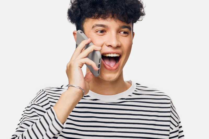 Young man talking on smartphone with excited expression, wearing striped shirt against white background, showing happiness and communication.