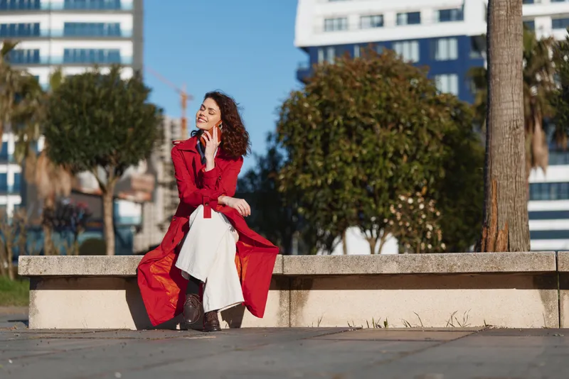 Young woman with curly hair enjoys a sunny day sitting on a concrete bench in a red coat and white skirt while talking on her phone outdoors.