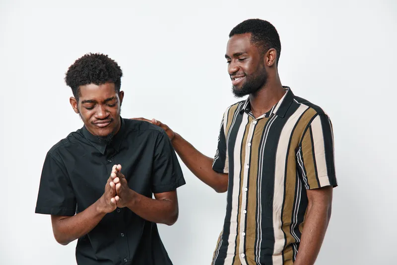 Two African American men with dark skin standing together on white background one comforting the other with hand on shoulder and supportive smile in casual clothing.