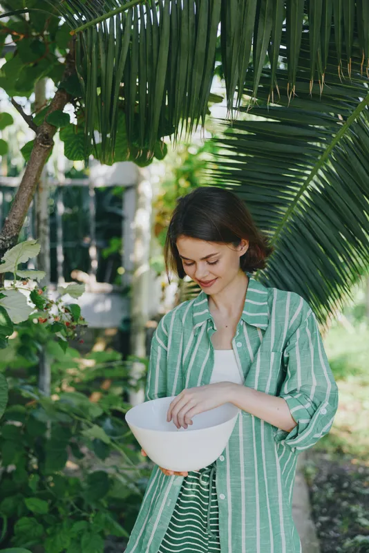 Woman in a lush garden holds a white bowl, wearing a green striped shirt. Outdoor light highlights a calm expression among leaves, creating a natural, peaceful mood in a candid portrait