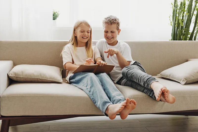 Two happy children sitting barefoot on a beige sofa reading a book together in a bright living room. Concept of childhood, learning, friendship, family bonding and relaxed home environment.
