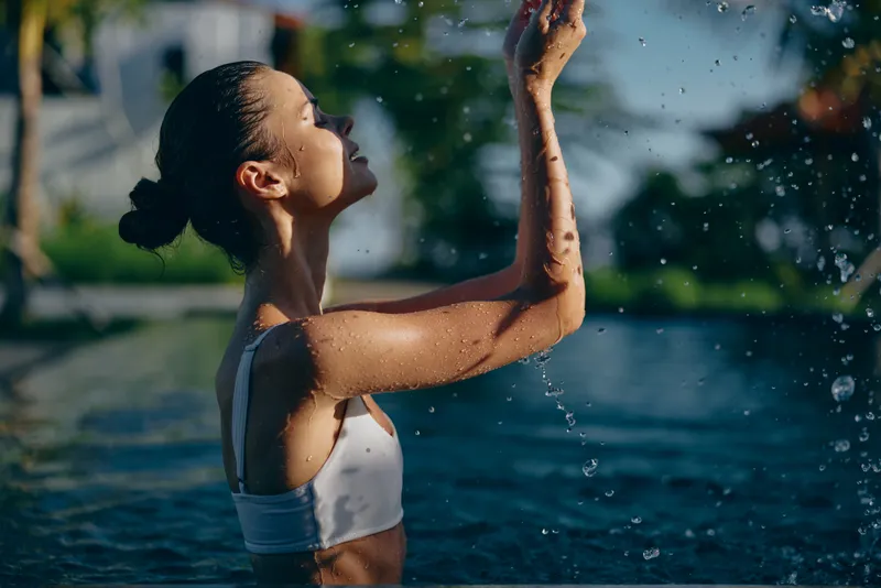 Joyful woman by a pool, water droplets suspended midair, sunlit outdoor scene, relaxed posture, white swimwear, vibrant greenery backdrop, casual lifestyle moment, fresh natural colors