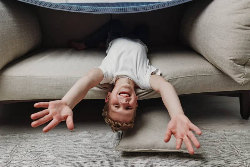 Happy boy lying upside down on sofa with arms stretched, smiling joyfully in cozy living room setting with natural light.