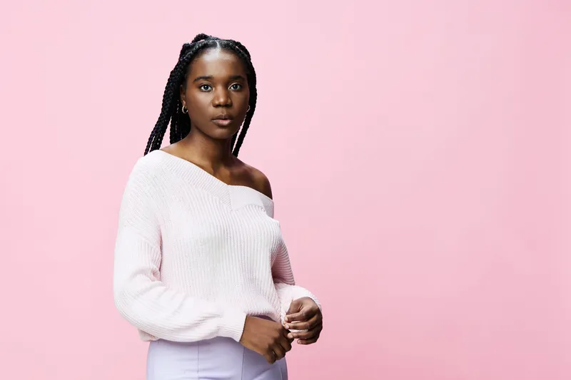 Elegant young black woman in a soft pink sweater poses against a pastel pink background, conveying confidence and style through her calm expression