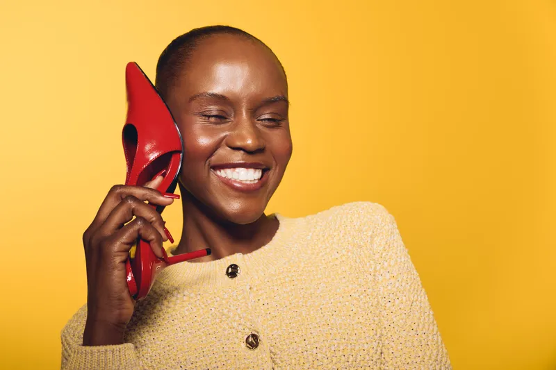 Smiling black woman with dark skin holds red high heel shoe near ear like phone, wearing beige sweater on yellow background. Concept of joy, fashion, and playful communication.