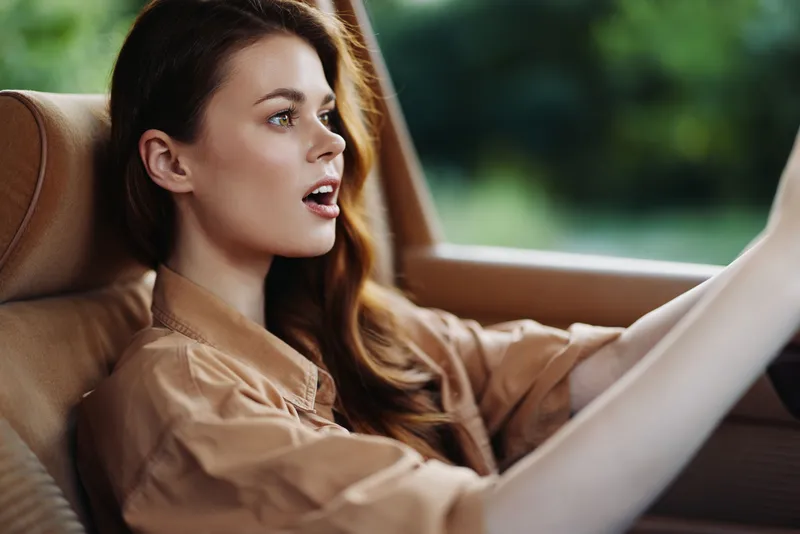 Young woman driving a car, expressing excitement and joy, captured in soft natural light with green background accents, showcasing casual fashion style.