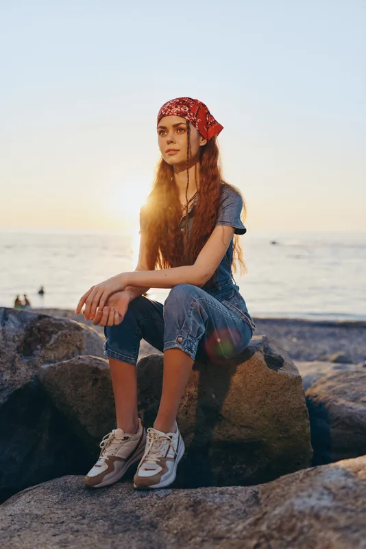 lifestyle woman in modern boho-western denim street style sitting on rocks by the sea during golden hour, film color with warm natural light and relaxed mood