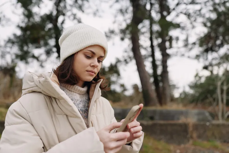 Woman in a warm beige coat and white beanie uses a smartphone outdoors in a park, winter light, focused expression, casual outfit, preparing messages amid bare trees and calm cold weather.