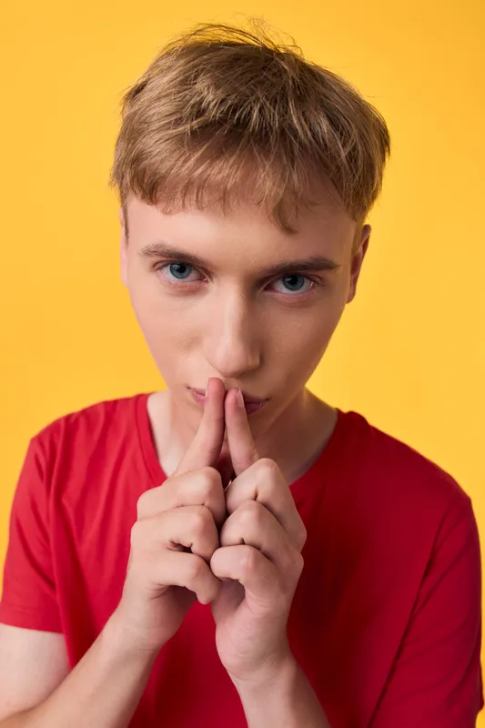 Silence, gesture, hush, youth, focus, introspection, contemplation, studio, closeup, calm illustration of a young man in a red shirt making a hush gesture against a vivid yellow background, conveying