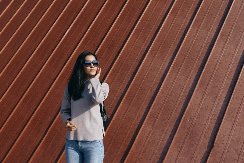 Woman wearing sunglasses in a casual sweater and jeans stands beside a diagonal wall outdoors, showcasing urban lifestyle and fashion with a confident pose, relaxed stance, street style vibe and