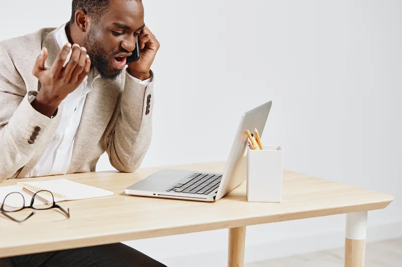 Young African-American man in beige blazer talking on mobile phone while sitting at a desk with laptop, glasses, and stationery, office workspace, casual business environment, indoor shot, modern tech