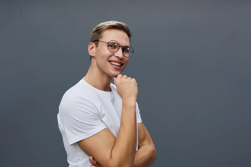 Smiling young man with glasses wearing casual white t-shirt posing against solid gray background, expressing happiness and confidence in studio portrait. People lifestyle concept, positive emotions, c
