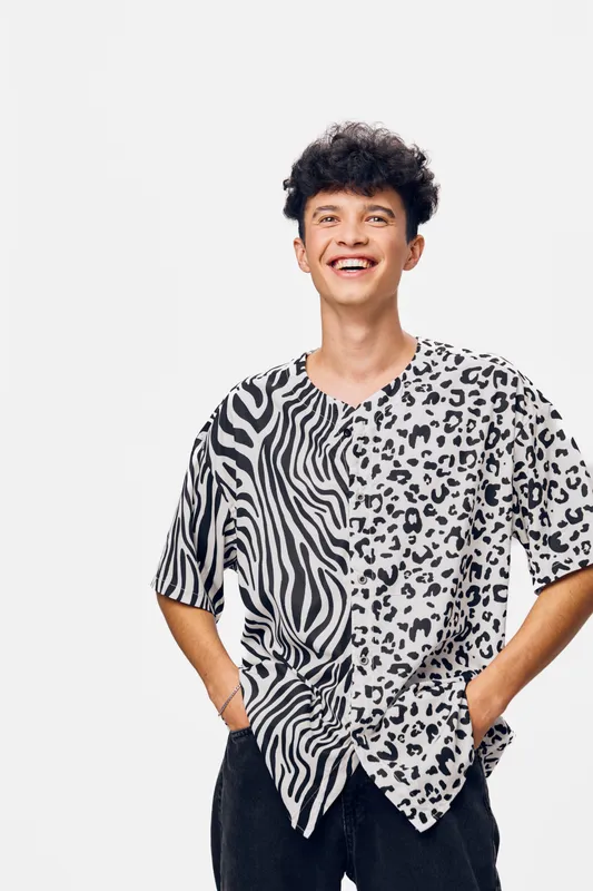 Young man smiling joyfully wearing a black and white animal print shirt with curly hair, standing relaxed on a plain white background, casual style and positive mood.