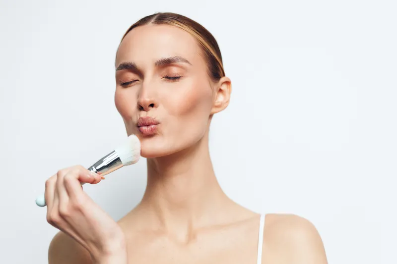 Young woman with closed eyes applying makeup with a brush, soft skin texture, natural beauty, minimalistic background, delicate expression, skincare routine concept.