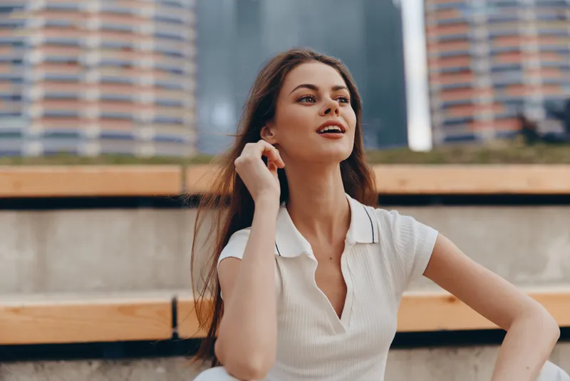 Young woman with long brown hair in white polo shirt smiling outdoors with urban background, casual fashion, positive expression, spring season