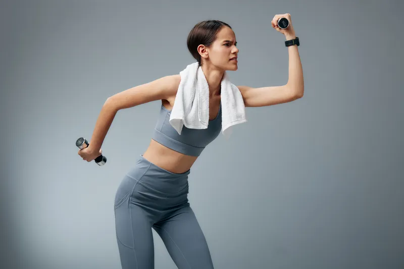 Athletic woman in gray activewear performs a dynamic dumbbell exercise with a towel draped over her shoulders in a clean studio.