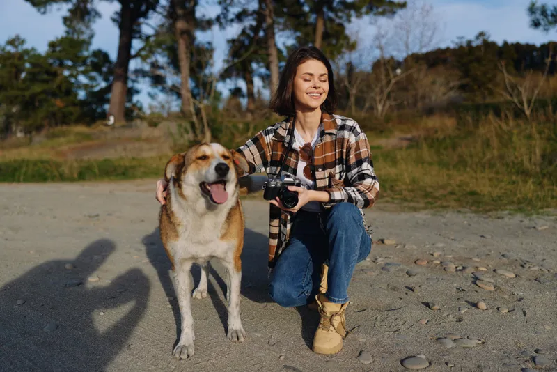 Woman with camera beside a playful dog on a sandy path, capturing candid moments in nature and enjoying a relaxed outdoor lifestyle together, under clear skies and gentle breeze.