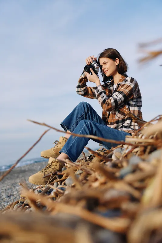 Female photographer crouches on sandy dunes with a camera, wearing a plaid shirt and jeans, capturing a coastal landscape with natural light and color.