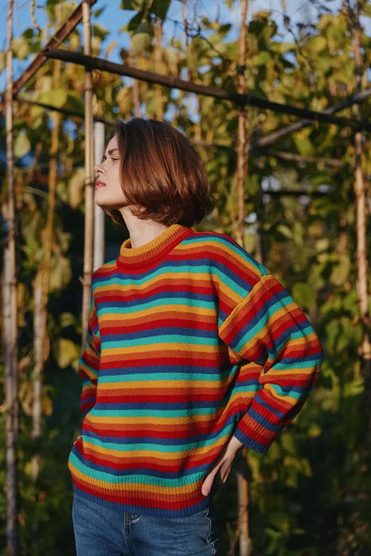 Woman in her mid 20s wearing a colorful striped sweater and jeans with a bob haircut, standing outdoors by a wooden gardening trellis in warm sunlight, hands on hip, serene lifestyle portrait.