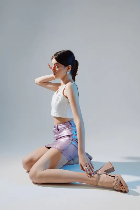 Young woman in a white top and shiny skirt poses kneeling on the floor wearing sunglasses. Studio shot with soft lighting. Concept for woman beauty fashion and style.