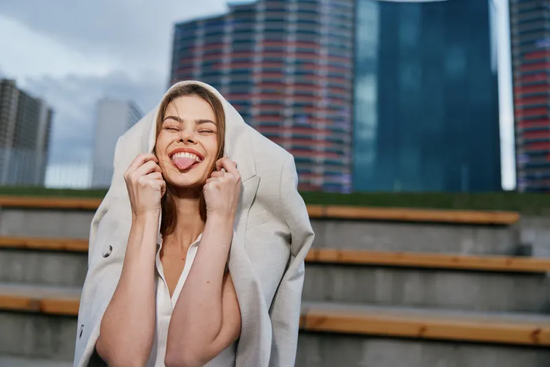 Playful woman showing tongue outdoors with coat on head in urban business environment, smiling confidently, casual white blouse, modern office buildings background