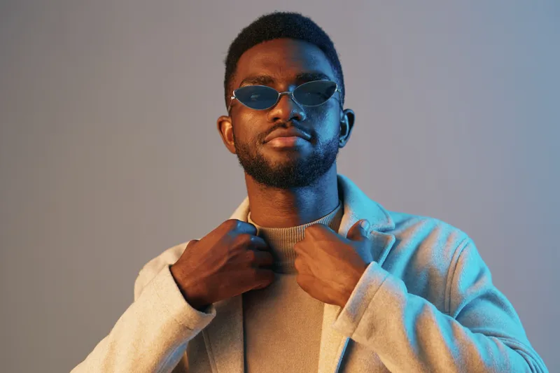 Stylish young man wearing sunglasses and a light coat, exuding confidence against a soft blue and orange background, ideal for fashion and lifestyle concepts