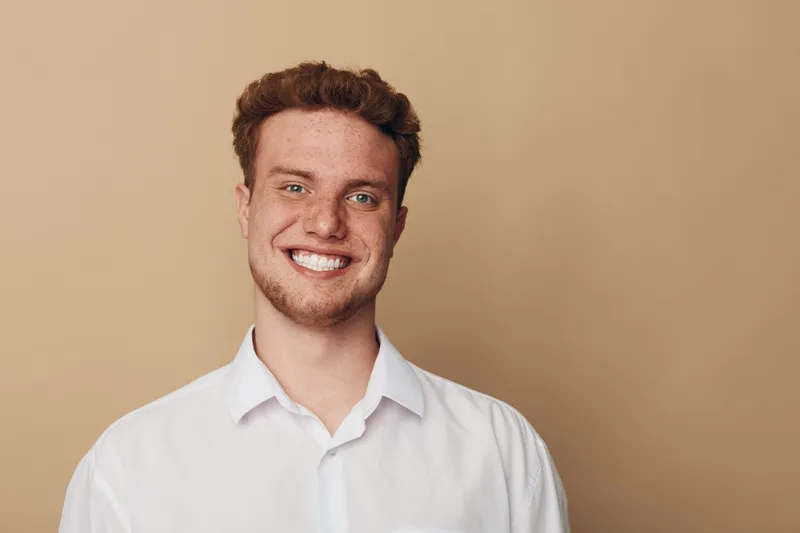 Smiling young man with curly hair and freckles wears white shirt against beige background. Portrait of happy confident male model with natural expression and casual style in studio lighting.