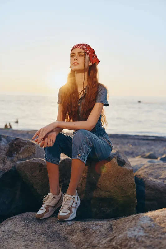 young woman with red bandana sitting on rocks at sunset by the ocean in casual blue outfit and sneakers, peaceful and relaxed mood, golden sunlight