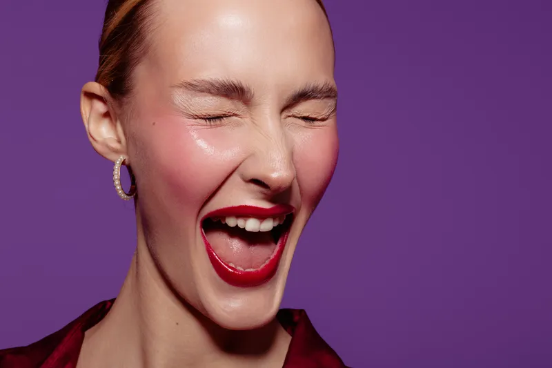 Close-up of a joyful woman with red lipstick and hoop earrings laughing with eyes closed on purple background. Concept of happiness, beauty, and vibrant emotions.