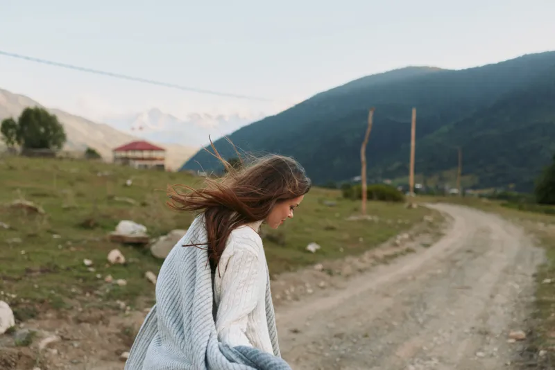Woman with windswept hair walking along a dirt road in a mountainous landscape, embodying outdoor lifestyle and calm wellness, soft natural light.