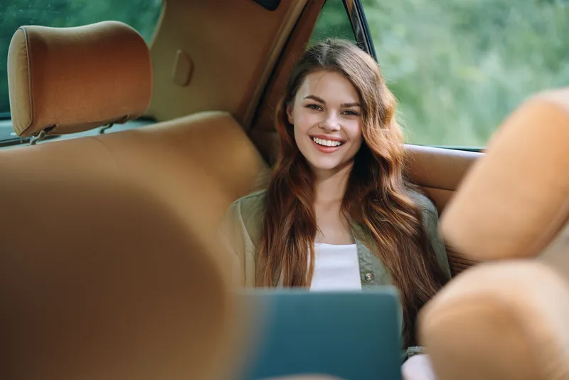 Smiling young woman working on a laptop in a car, enjoying a relaxed moment in nature with a vibrant background and warm color palette.