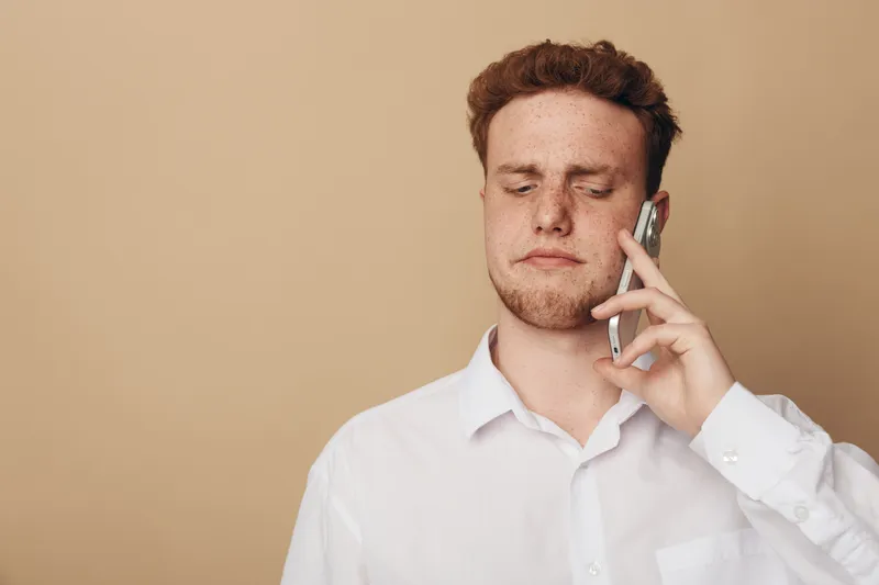 Young man in white shirt holds smartphone to ear with eyes closed, showing concentration or frustration. Neutral beige background emphasizes emotion and communication concept.