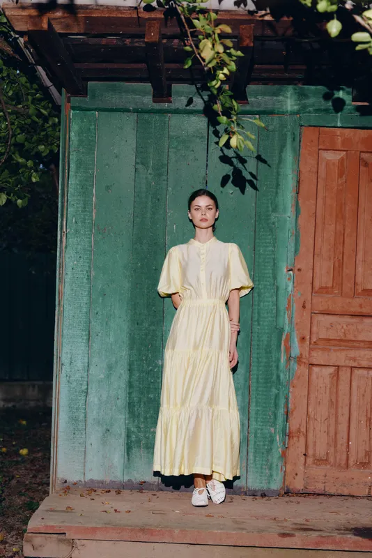 Fashionable woman in a pale yellow dress stands before a weathered green wall and orange door, creating a serene rustic portrait with soft natural light.