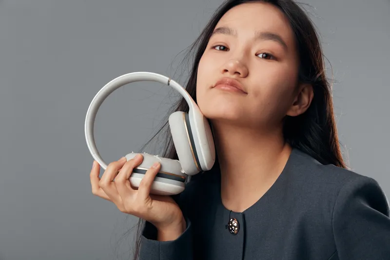 Young woman holding headphones close to face with confident expression in studio setting. Portrait of female with long dark hair wearing dark jacket for music or technology concept.