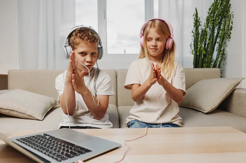 Two children in casual clothes wear headphones and clap hands in living room with laptop on wooden table. Concept of playful learning, music enjoyment, and modern technology use at home.