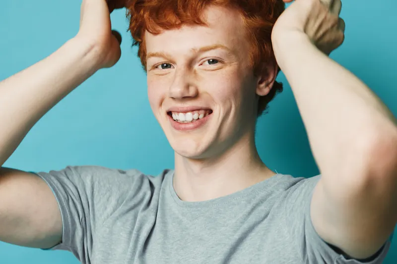 Young man with red hair and freckles smiling with hands near head on blue background. Casual style in gray shirt with happy smile and joyful emotion in studio portrait.