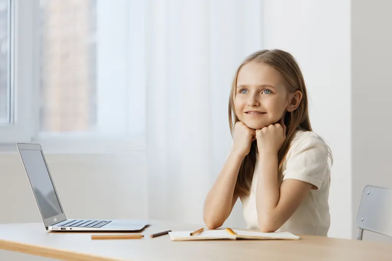 Thoughtful young girl contemplating at a desk with a laptop, exuding joy and curiosity in a bright, minimalistic home office setting