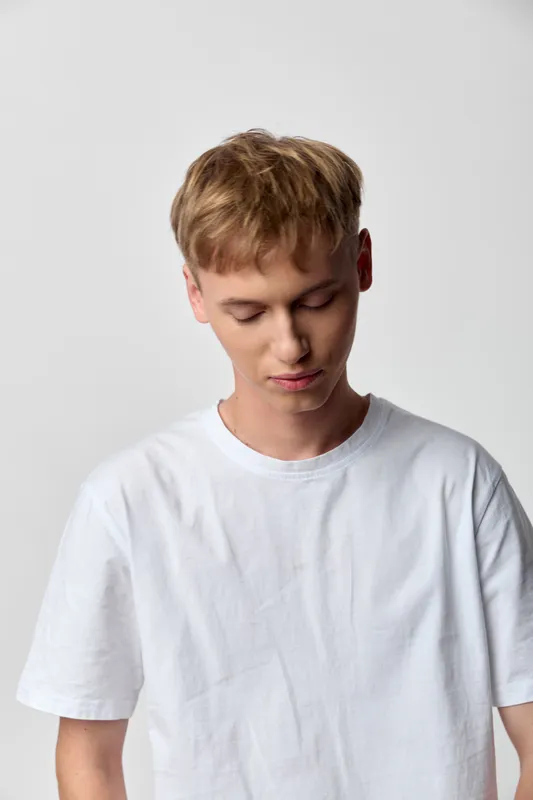 Young man in a white tee gazing downward in a minimal studio setting, casual wear and calm mood, focused on inner thought, soft lighting highlighting clean fabric and simple style.