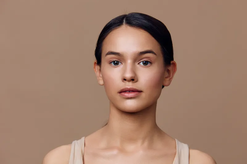 Portrait of a young woman with a neutral expression, featuring a natural beauty look against a warm, minimalist background emphasizing skin tones and simplicity