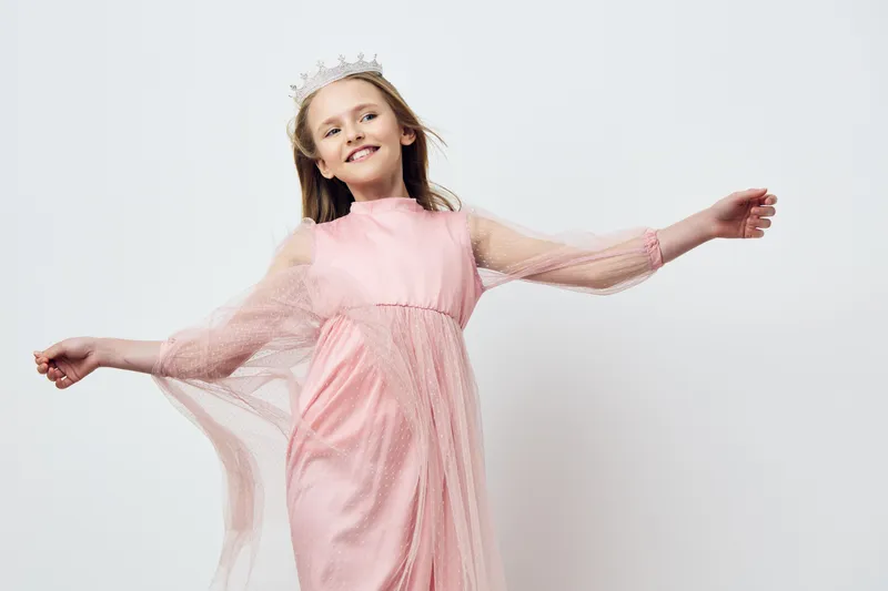 A joyful young girl in a pink dress with flowing sleeves and a crown, dancing gracefully against a minimalist white background, embodying innocence and playful childhood