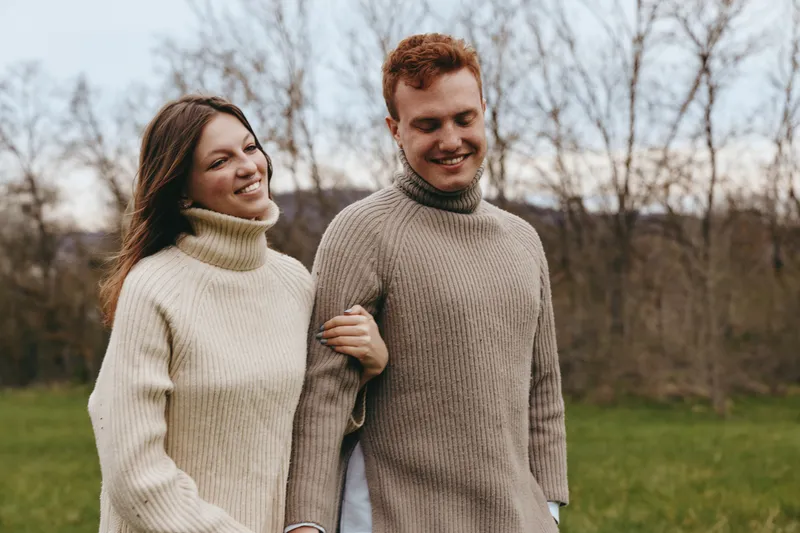 Young couple in knitted sweaters walk arm in arm outdoors in late autumn enjoying a peaceful moment together with soft natural light and bare trees in the background