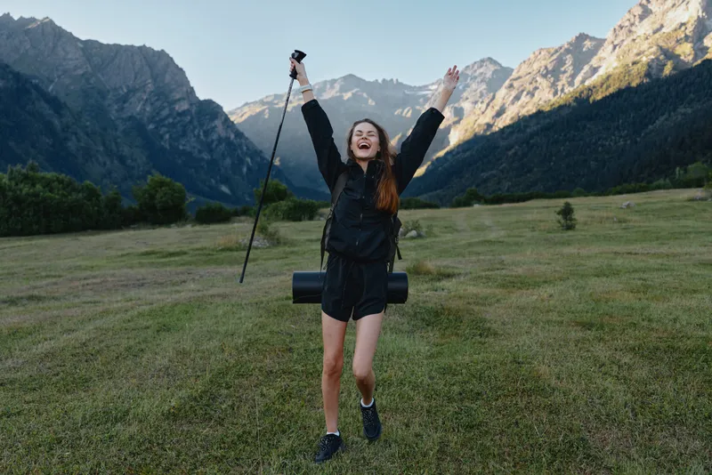 A joyful female hiker stands in a wide valley with towering mountains, arms raised in triumph after a trek, wearing outdoor gear and a backpack, radiating satisfaction and energy.