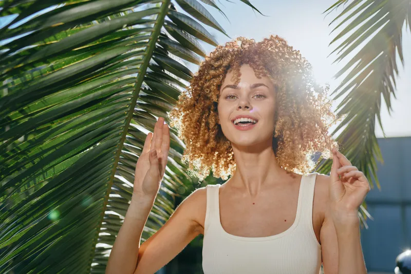 smiling woman with curly hair enjoys sunny tropical outdoors near palm leaves, wearing white tank top and expressing joy and freedom during daytime.